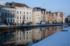 people walking on the side of a river in front of old buildings and snow covered ground