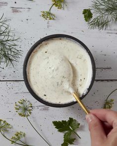 a hand holding a spoon in a bowl filled with ranch dressing surrounded by flowers and herbs