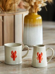 two mugs with red crabs on them sitting on a table next to a vase