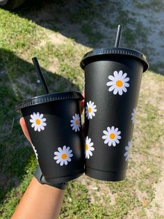 two black cups with white and yellow daisies on them are being held by someone's hand