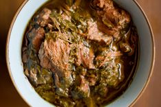 a white bowl filled with meat and green sauce on top of a wooden table next to a spoon