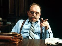 an older man with glasses and suspenders sitting in front of a table holding a book