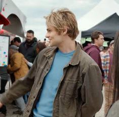 a young man standing in front of a group of people at an outdoor event wearing a blue shirt and brown jacket