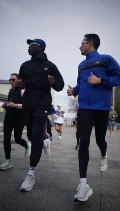 two men are running in the street with other people behind them wearing blue jackets and black pants