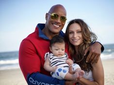 a man holding a baby on the beach with his arm around a woman's shoulder