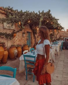 a woman standing in front of tables with chairs and potted plants on the wall