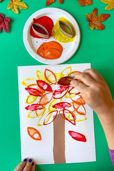 a child's hands painting a tree on paper with autumn leaves around the base