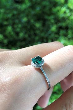 a woman's hand holding a ring with a green stone on it, in front of some bushes