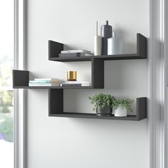 two black shelves with plants and books on them in front of a white wall next to a window