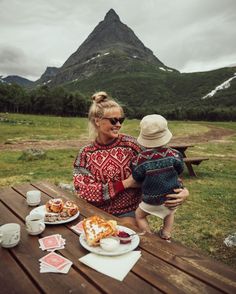 a woman holding a child while sitting at a picnic table in front of a mountain