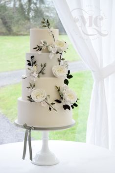 a three tiered white wedding cake with flowers on the top and sides, sitting in front of a window