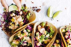 three tacos with various toppings are on a white surface next to lime wedges