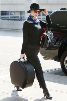 a woman in black is carrying a suit case and hat while walking down the street