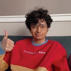 a young man with glasses sitting on a couch giving the thumbs up sign while wearing a red, yellow and blue shirt