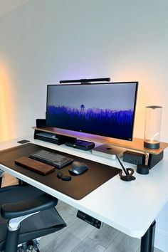 Modern desk setup featuring a large monitor with a serene forest wallpaper, complemented by a backlight that casts a soft glow on the white walls. The workspace includes a wooden monitor stand, a black keyboard, and various tech accessories, all arranged on a clean white desk for a streamlined look. Desk Setup Workspace Inspiration, Desk Setup Workspace, Gaming Computer Room, Minimal Workspace, Coding Tutorials