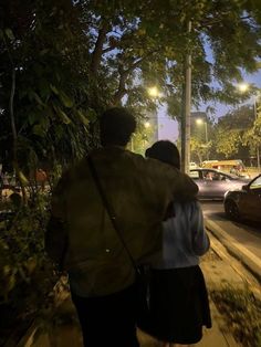 two people walking down the street at night with cars parked on the side of the road