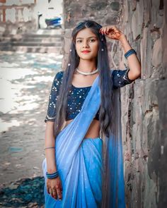 a woman with long hair wearing a blue sari and standing next to a brick wall