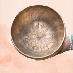 a person's hand holding a metal bowl with an intricate design on it, against a pink background