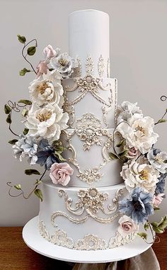 a three tiered white wedding cake with flowers on the top and sides, sitting on a wooden table