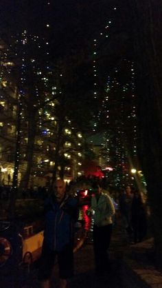 two people are standing in the dark near some buildings and trees with lights on them