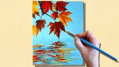 a person is holding a paintbrush and painting leaves on a piece of paper with water in the background