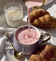 a cup of pink liquid being poured into a saucer next to croissants