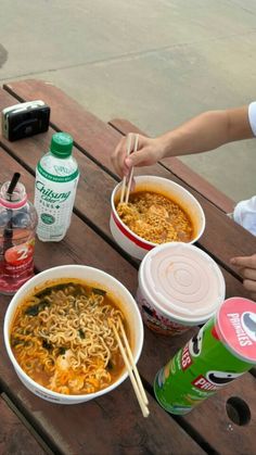 two bowls of ramen are sitting on a picnic table with drinks and sodas