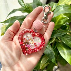 a hand holding a heart shaped keychain in front of a potted plant