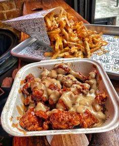 some food is in a white container on a wooden table and fries are next to it