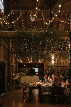 people sitting at tables in a barn with lights strung from the ceiling and greenery