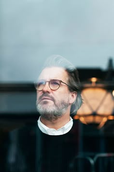 a man with glasses and a beard looking at something in the distance through a glass window