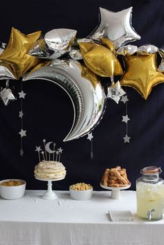 a table topped with balloons and stars next to desserts on top of a white table cloth