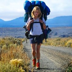 a woman walking down a dirt road with backpacks on her back