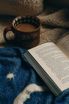 an open book sitting on top of a blanket next to a cup of hot chocolate