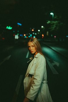 a woman standing on the side of a road at night with her hands in her pockets