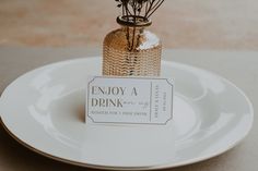 a small bottle with flowers in it sitting on a plate that says enjoy a drink