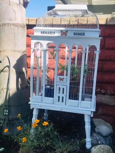 a white birdcage sitting on top of a flower bed next to a brick wall