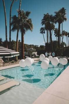 some white balloons floating in the air near a swimming pool with palm trees and chairs