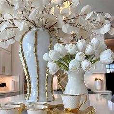 white and gold vases with flowers in them on a countertop next to cups