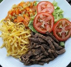 a white plate topped with pasta, meat and vegetables next to tomatoes on top of lettuce