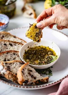 a person dipping some bread into a small bowl with pesto on it and other food in the background