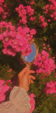 a person's hand holding a mirror in front of pink flowers