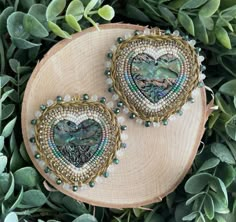 two heart shaped brooches sitting on top of a piece of wood next to green leaves