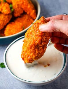 a person dipping something into a bowl with sauce on it and chicken wings in the background