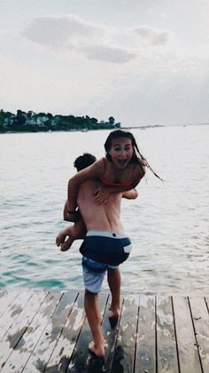 a man carrying a girl on his back while standing on a dock over water with houses in the background