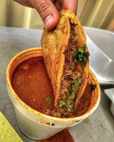 a hand dipping some food into a bowl filled with red sauce and garnished with cilantro