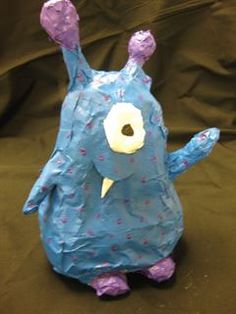 a blue paper mache monster sitting on top of a black cloth covered floor next to a brown wall