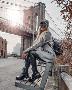 a woman sitting on a bench in front of the brooklyn bridge wearing black boots and a gray coat