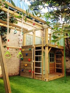 a wooden play structure with swings and climbing bars in the grass next to a brick wall