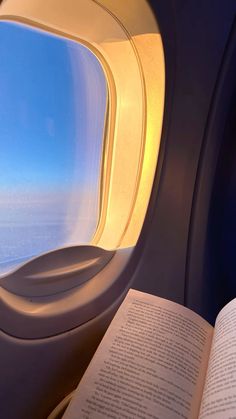 an open book sitting on top of an airplane seat next to a window with the sky in the background